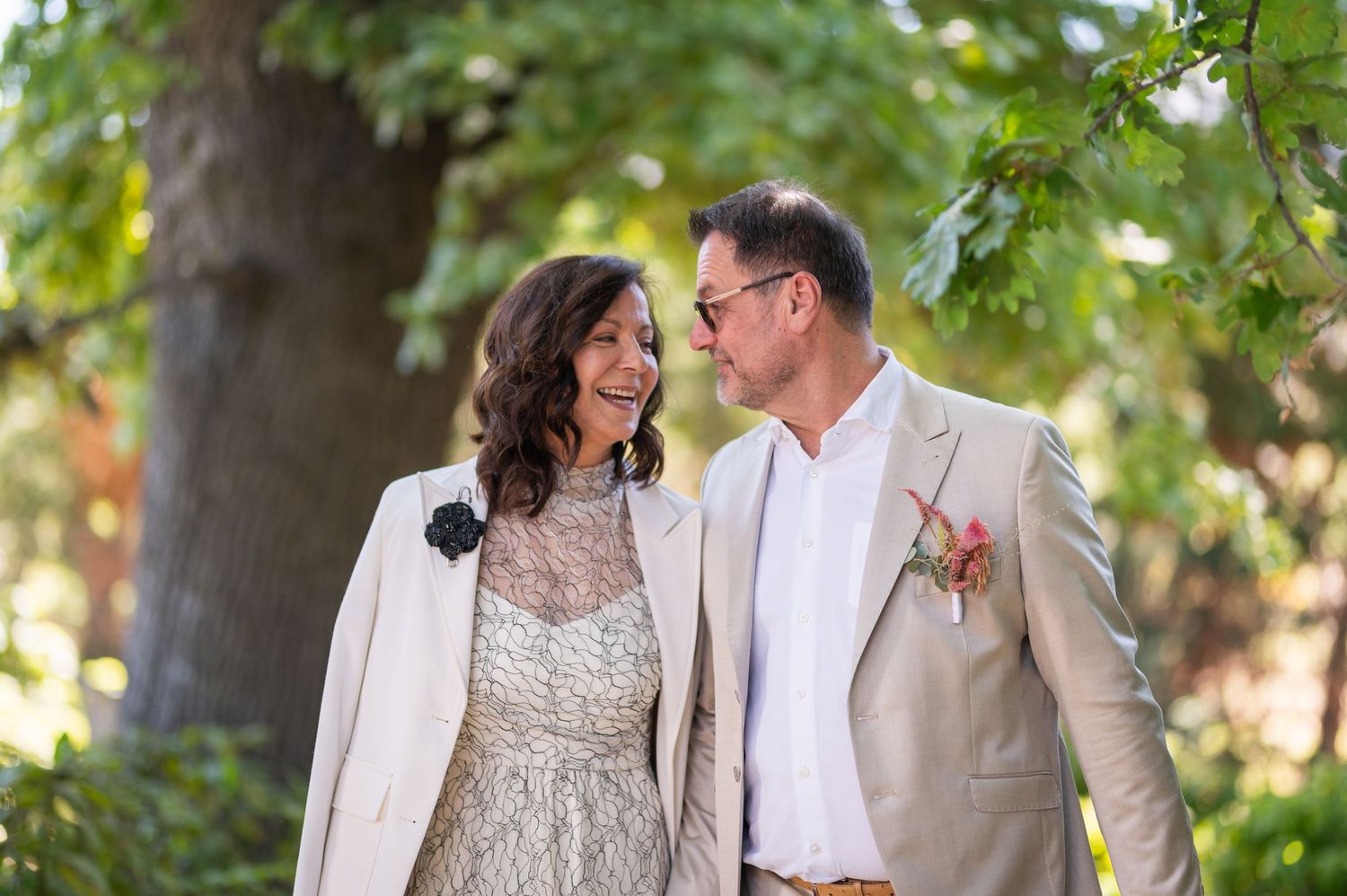 A smiling couple in elegant wedding attire stands outdoors under lush green trees, sharing a joyful moment together.