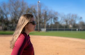 Person wearing sunglasses and a red jacket standing on a baseball diamond with bare trees in Charleston South Carolina