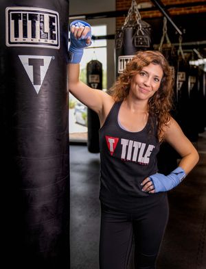 Boxer posing confidently beside a black Title punching bag wearing workout gear and blue hand wraps at Title Boxing Gym in Summerville, SC
