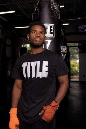 Person wearing black TITLE t-shirt and orange gloves poses in dark gym setting with dramatic lighting.