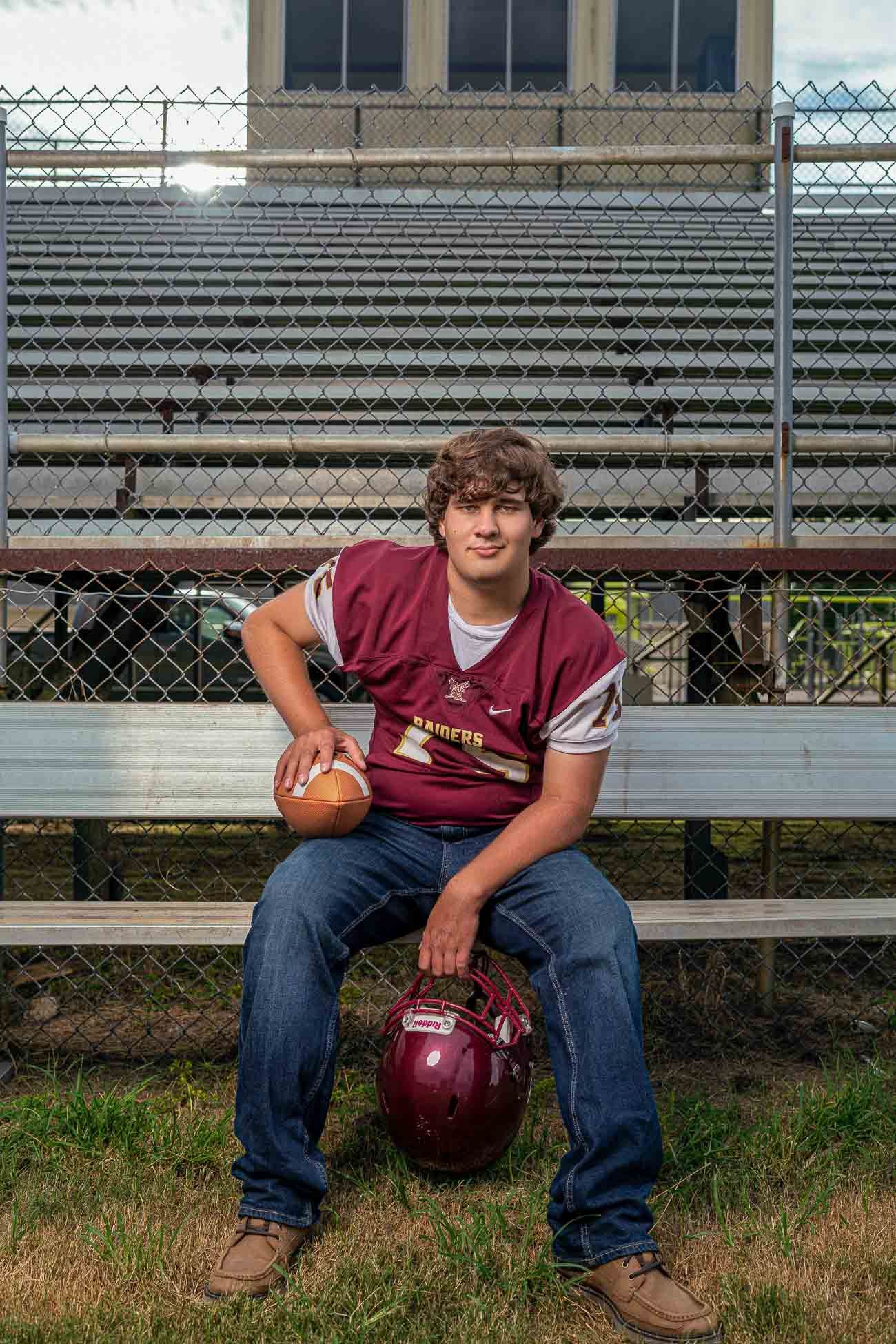 Dorchester Academy football player in maroon jersey sitting on metal bleachers holding a football