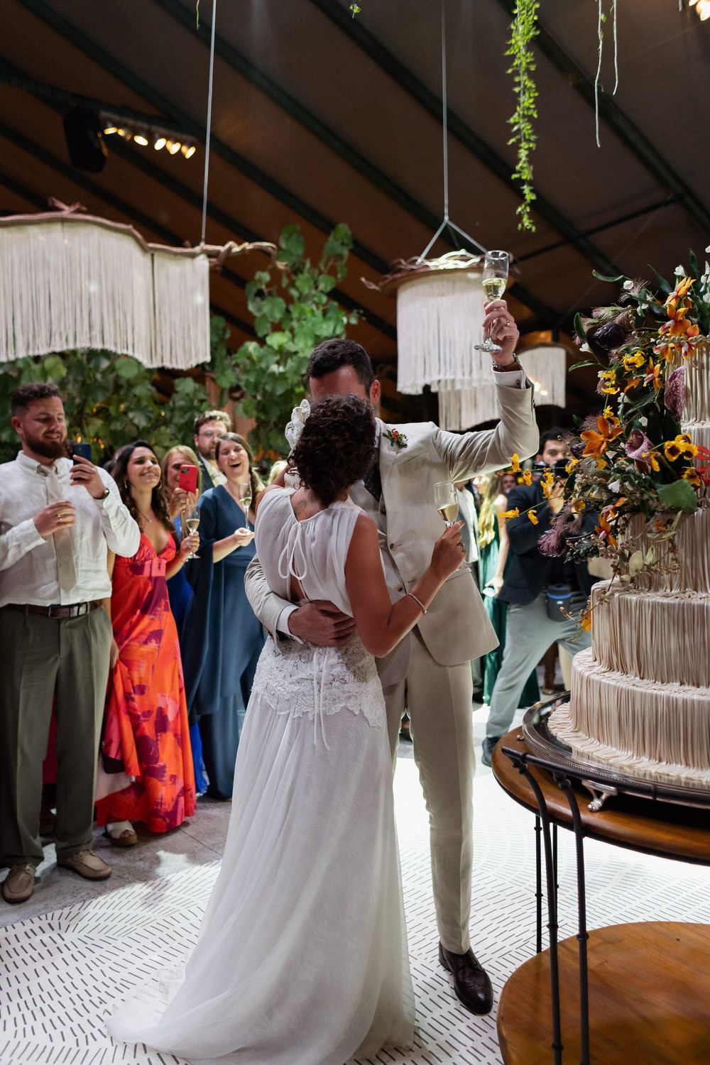 Wedding couple and guests gather around a tall white wedding cake during the cake cutting ceremony at an indoor venue.