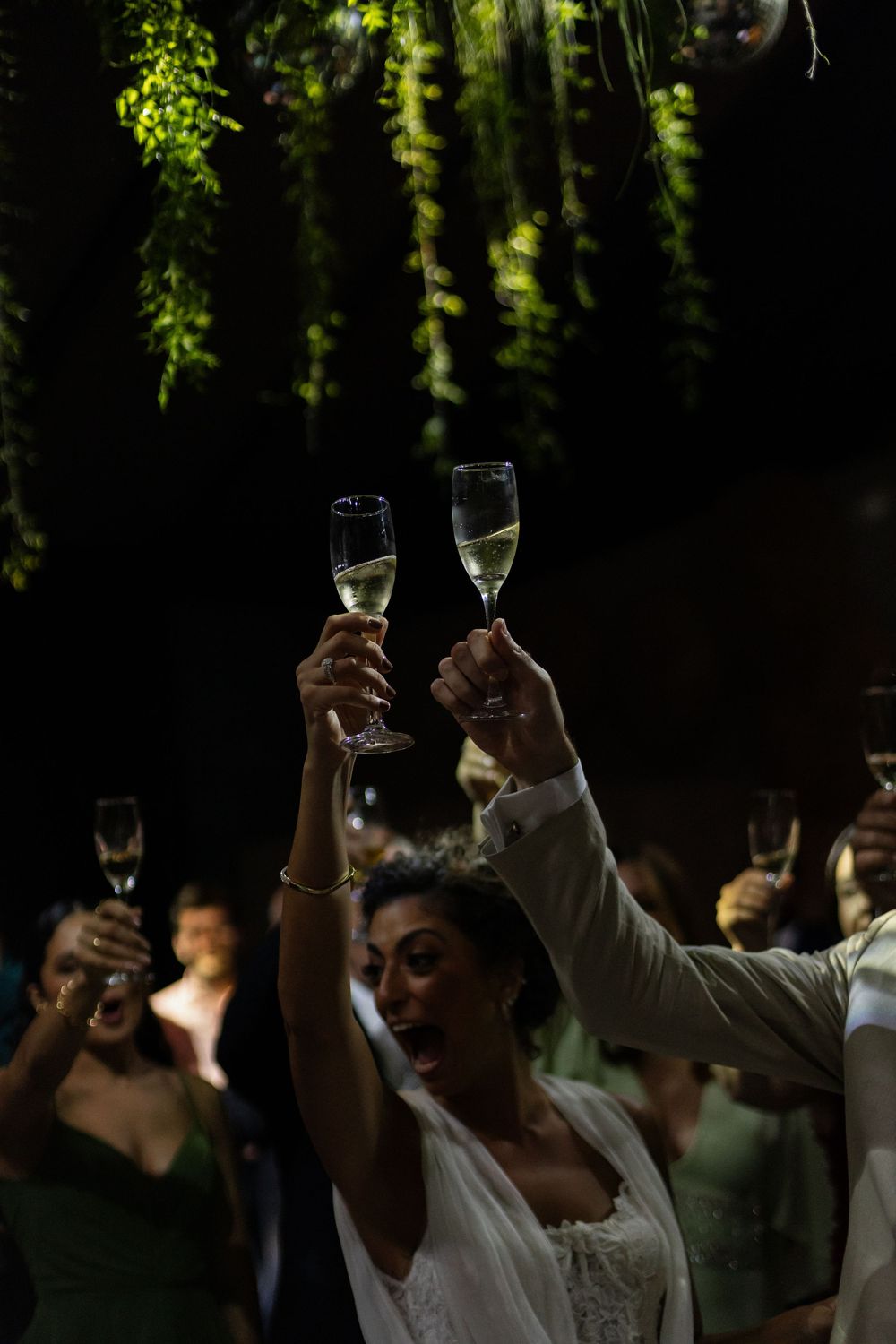 Wedding guests raising champagne glasses for a toast under hanging greenery and dim lighting.