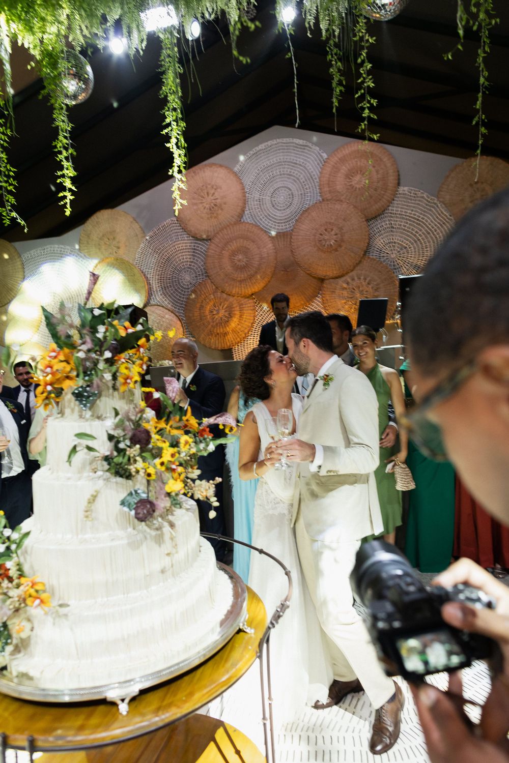An elegant white wedding cake displayed under hanging greenery and decorative lanterns.