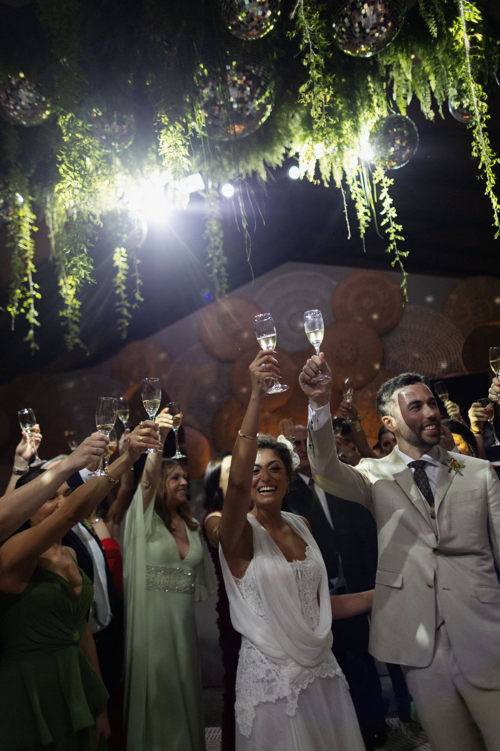 Wedding guests raise champagne glasses for a celebratory toast under twinkling lights and hanging greenery at night.