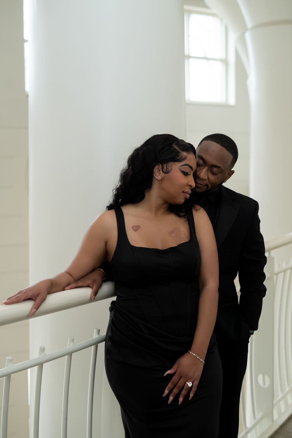 A romantic couple in black clothing embraces and kisses in a bright white hallway with windows.