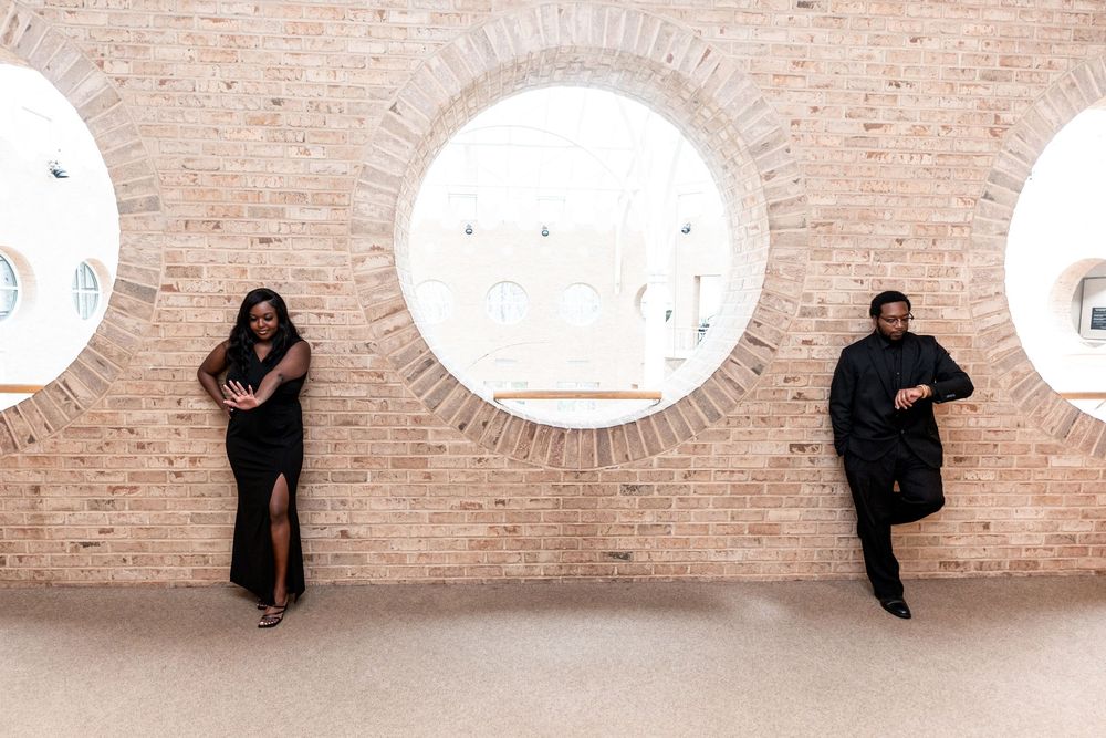 Two people pose creatively against a beige brick wall with circular window cutouts.