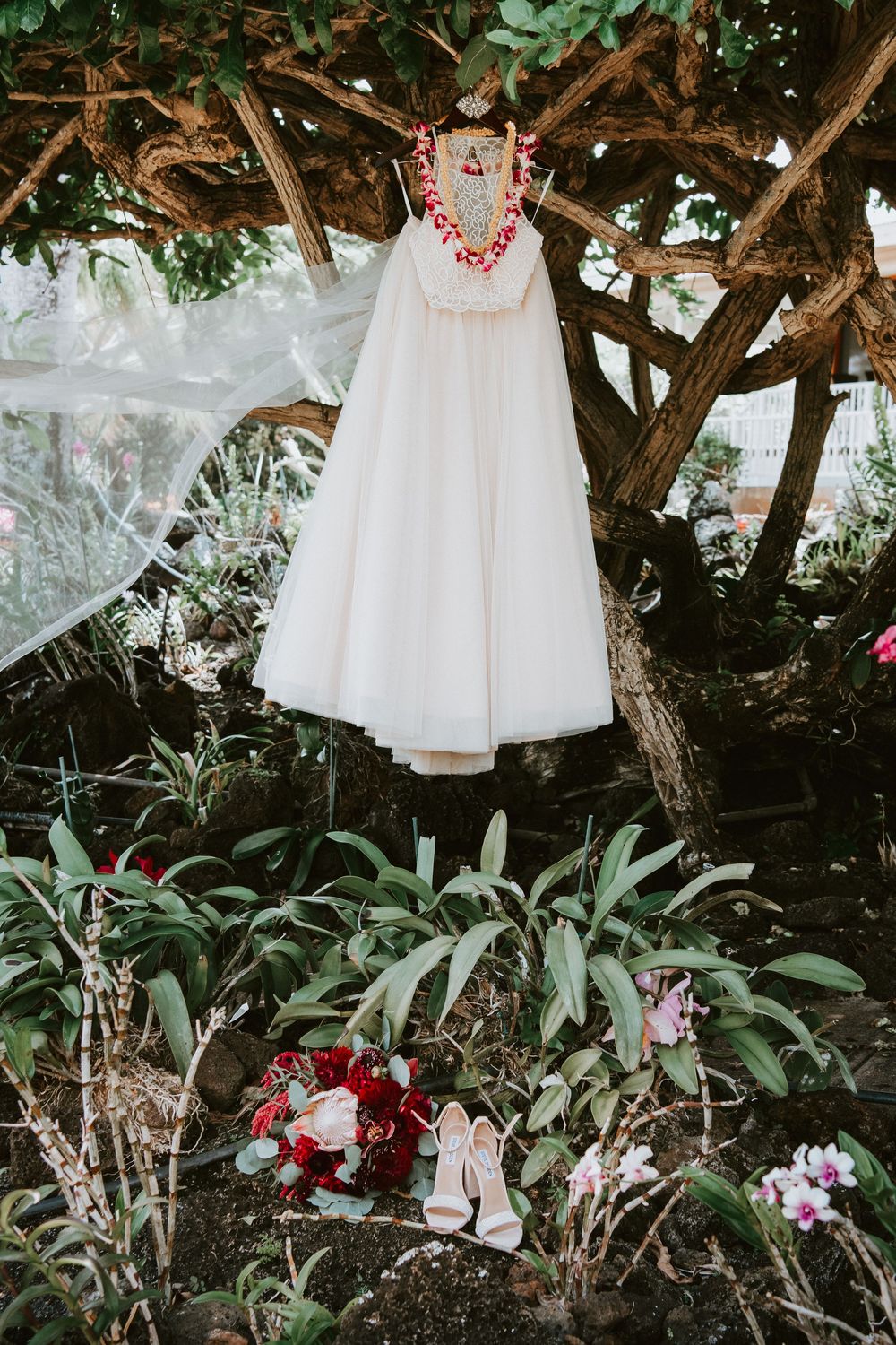 White halter wedding dress hanging from tropical tree with bridal heels and red bouquet below.