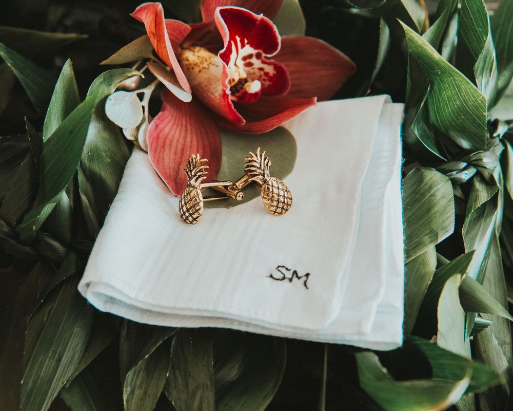 Gold pineapple cufflinks and monogrammed white handkerchief with red orchid on tropical green leaves.