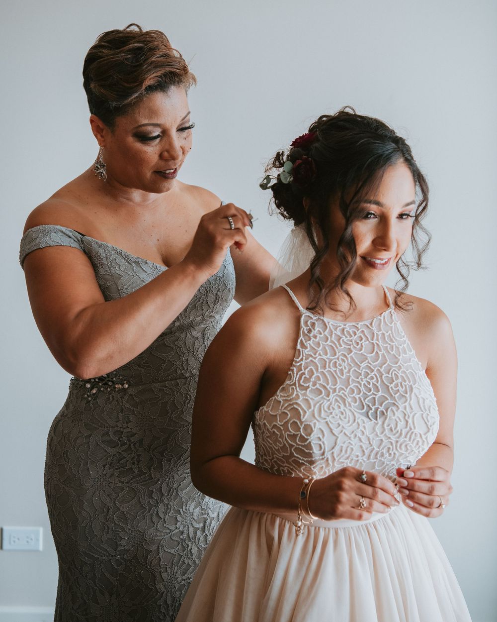Mother helps bride fasten her white lace halter wedding dress while bride smiles softly indoors.