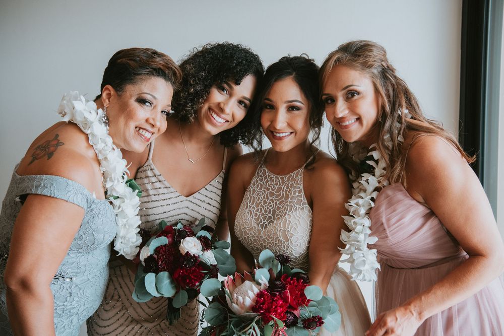 Bride and three bridesmaids in silver and pink dresses smile together wearing white floral leis.