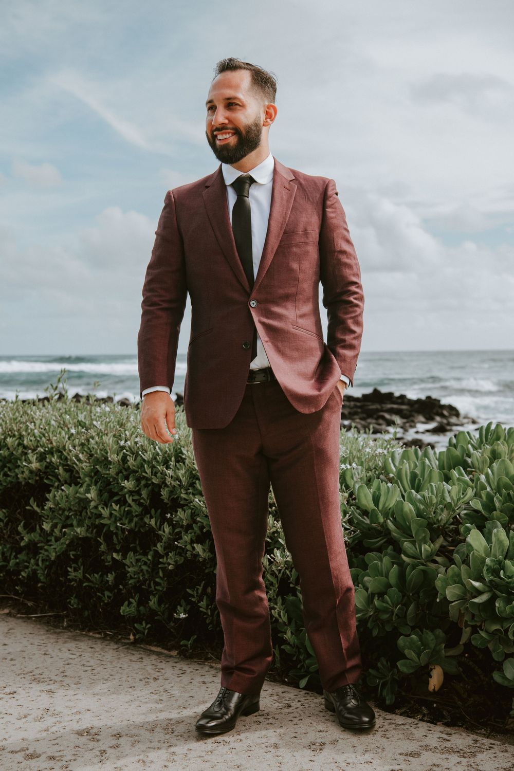 Handsome groom in burgundy suit stands confidently on beach path with ocean waves behind him.