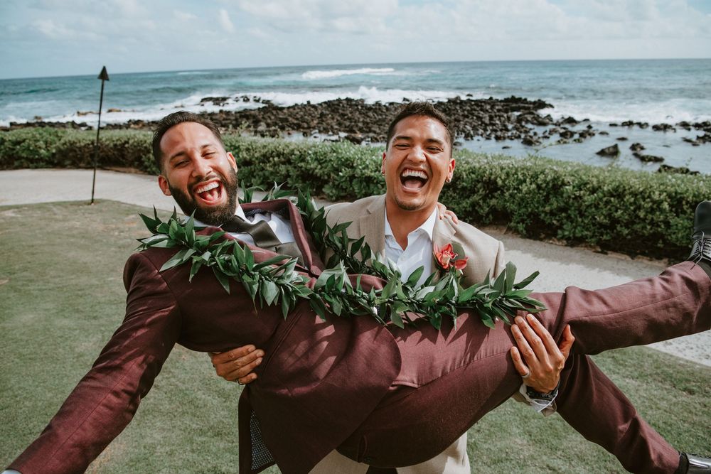 Two laughing groomsmen in burgundy and tan suits joke around with one being carried outdoors.