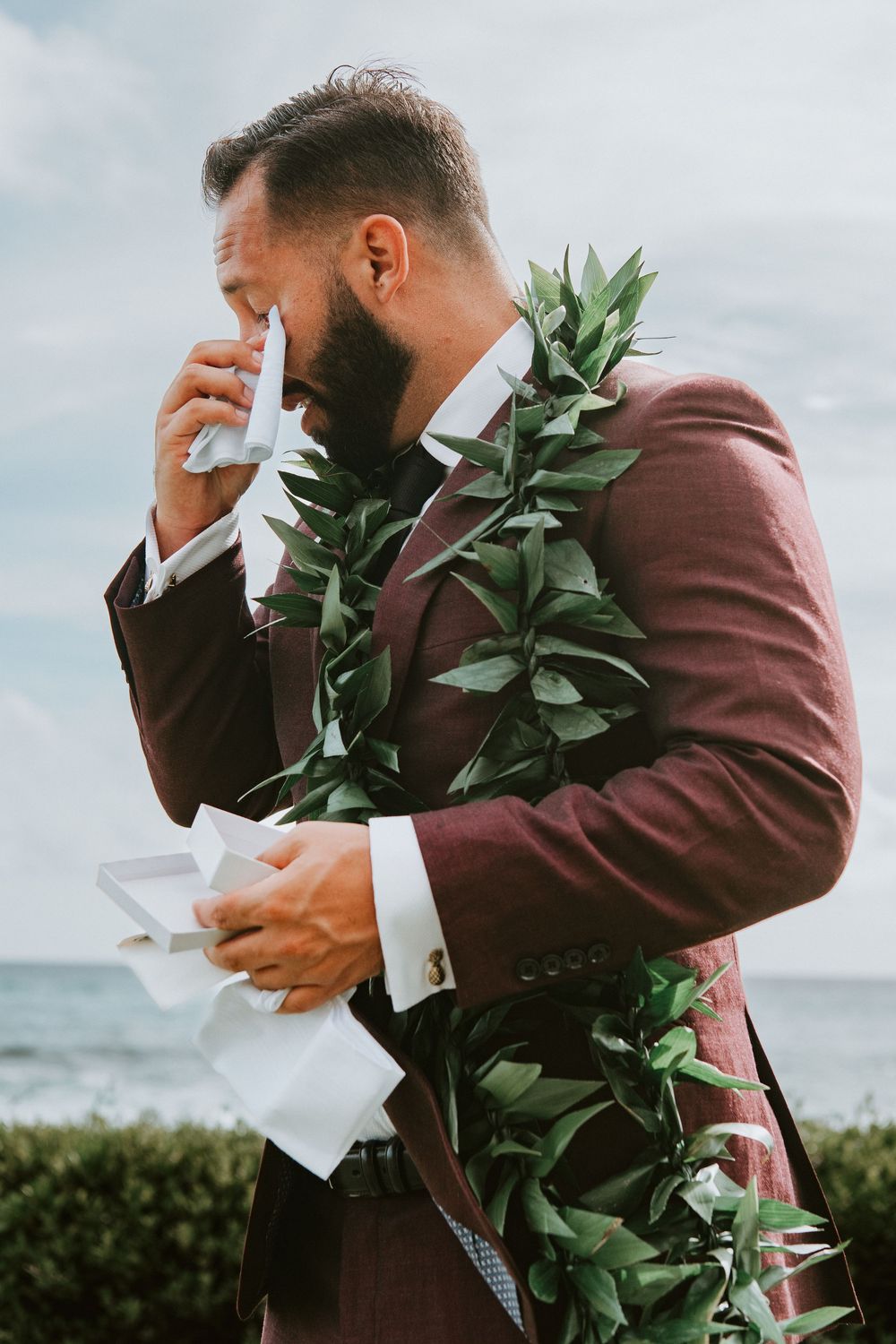 Emotional groom in burgundy suit with green leaf lei wiping tears at outdoor Hawaiian wedding ceremony.