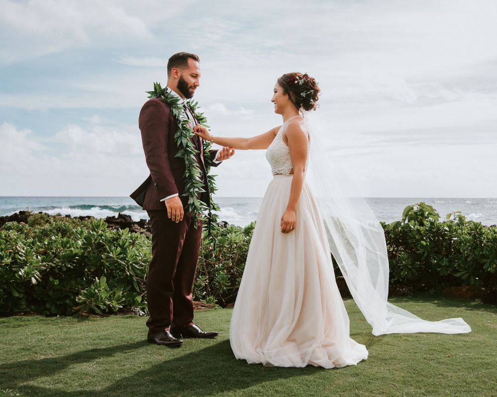 Bride in white gown and groom in burgundy suit exchange vows at oceanfront Hawaii wedding.