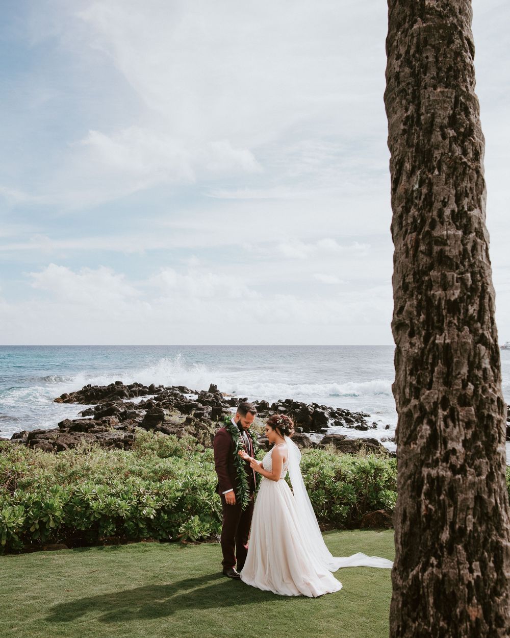 Bride and groom share intimate moment by rocky Hawaiian coastline framed by a tall palm tree.