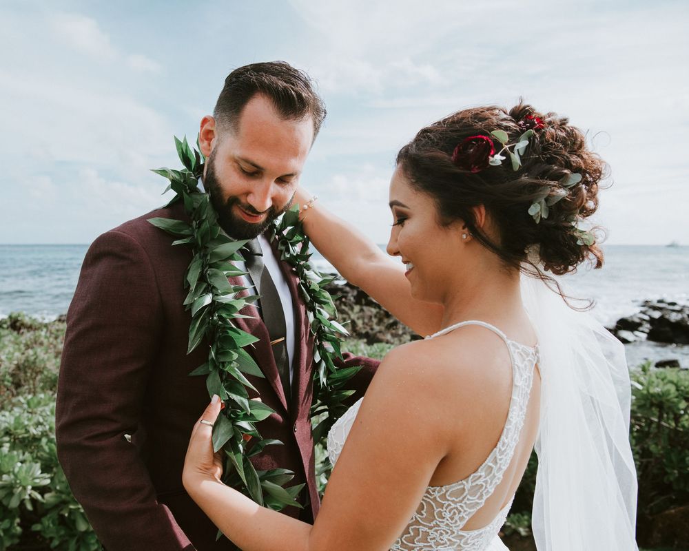 Bride placing green leaf lei on bearded groom in burgundy suit at Hawaii beach wedding.