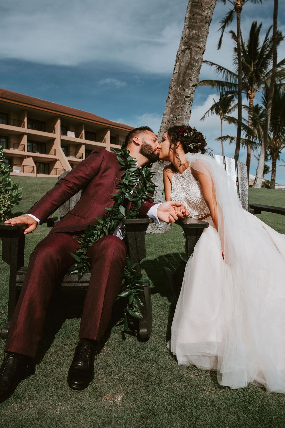 Bride and groom share a kiss while seated on benches with palm trees at Hawaii resort wedding.