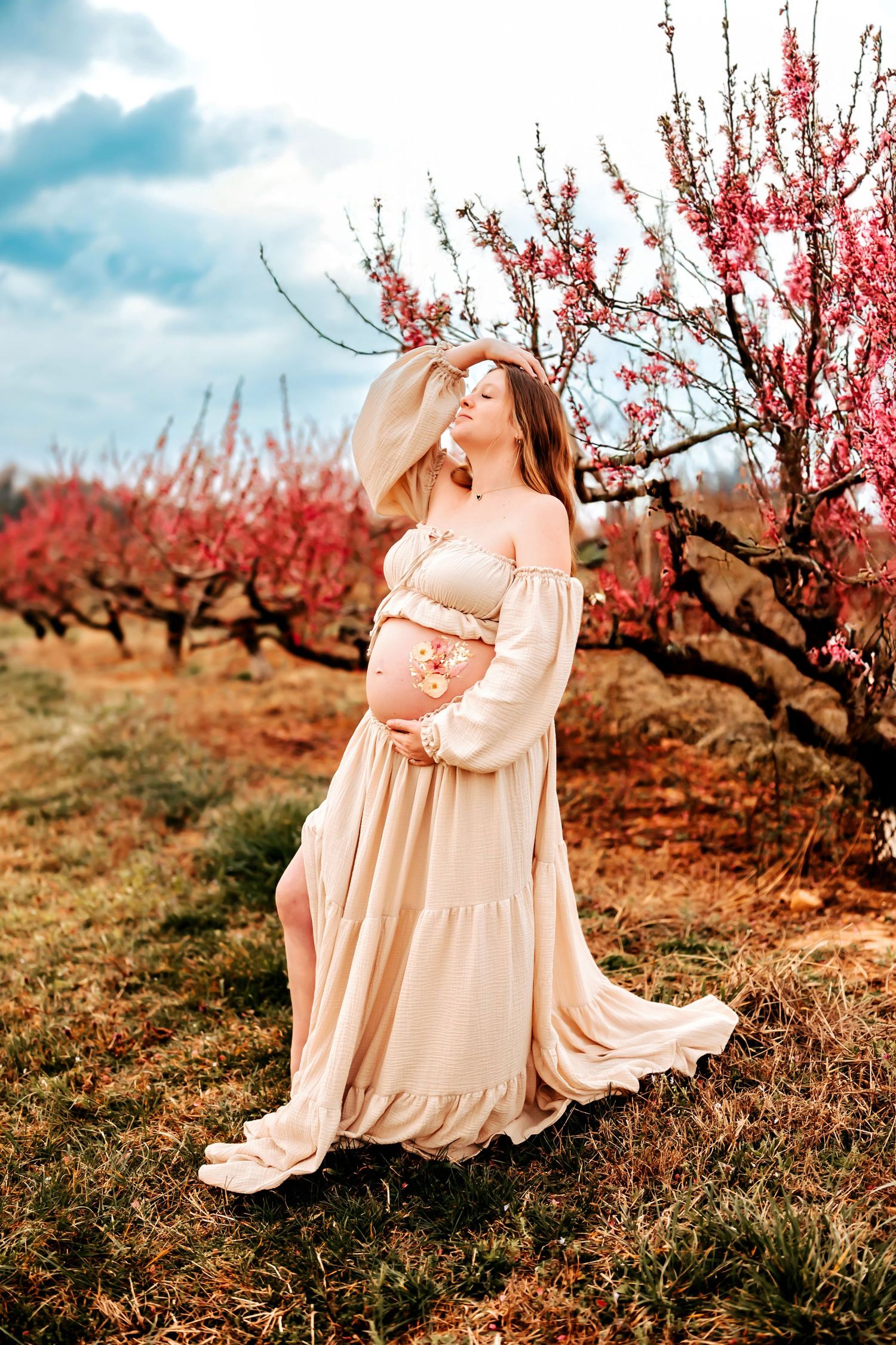 A figure in a flowing cream dress poses gracefully among blooming pink peach trees in a spring orchard setting.