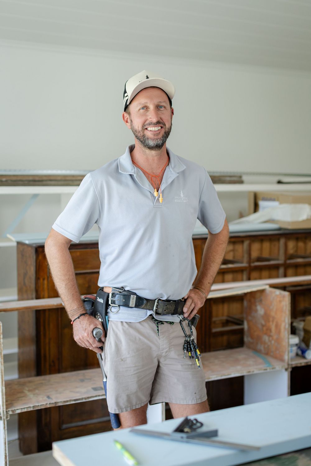 Contractor in blue polo and shorts holds power tools while working on home renovation project in workshop setting.