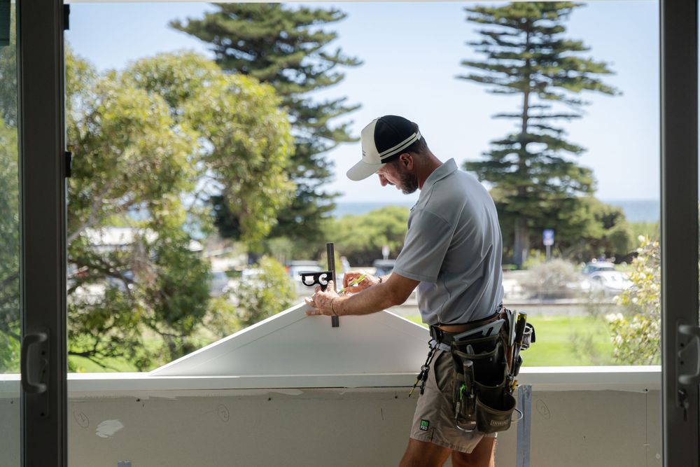 Carpenter working on a house with ocean and pine trees visible through balcony windows.