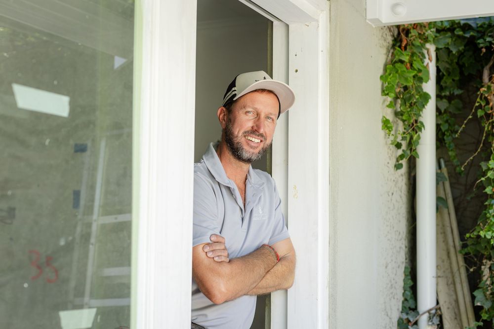 A tradie wearing a white cap leans against a white doorframe with crossed arms and a relaxed smile.