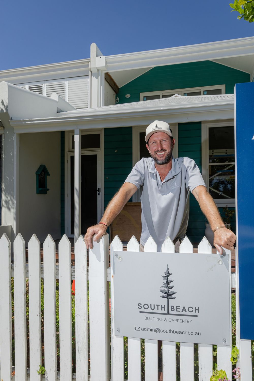 Business owner and builder stands behind a white picket fence with a South Beach Building signage on a sunny day.