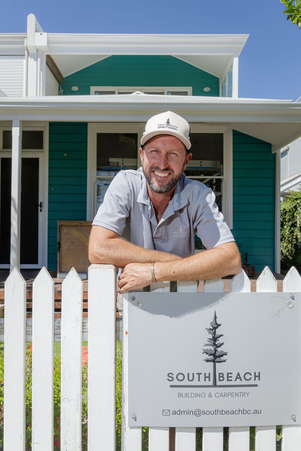 Carpenter smiling in white hat leaning over a white fence with South Beach Building & Company signage.