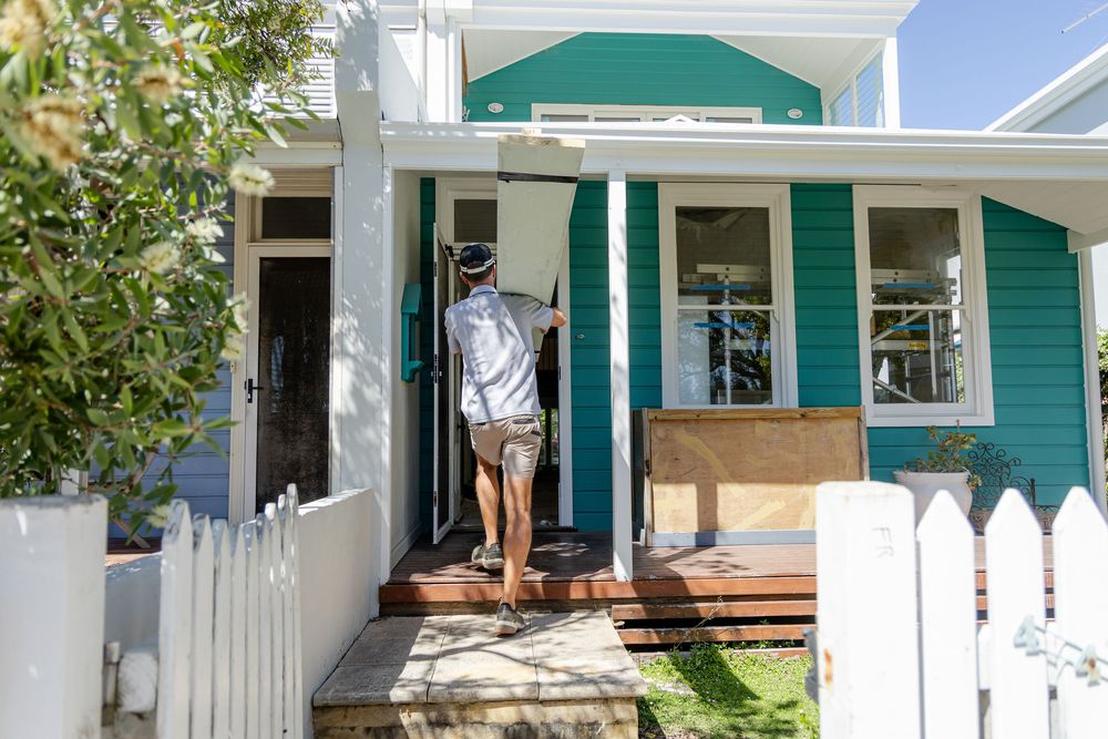 Branding photos of a builder in shorts walks up the steps of a teal-colored cottage with white trim and a picket fence carrying building mat