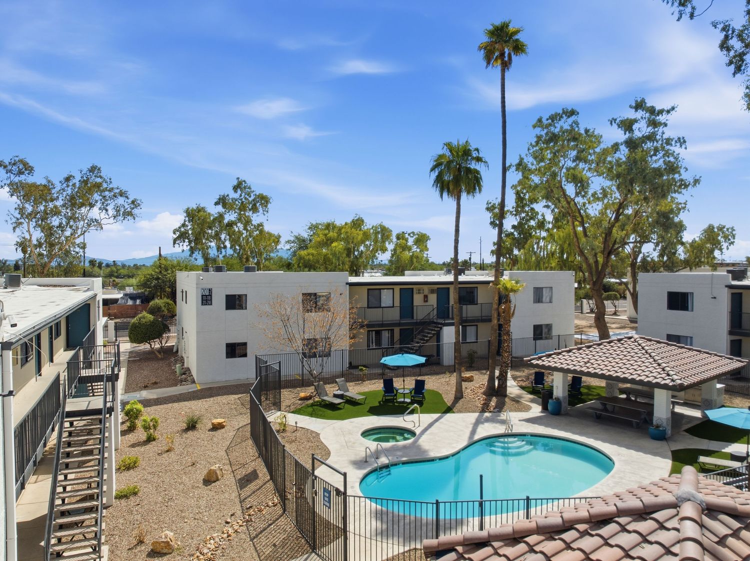 Modern apartment complex exterior with swimming pool and palm trees under blue sky in a sunny desert setting.