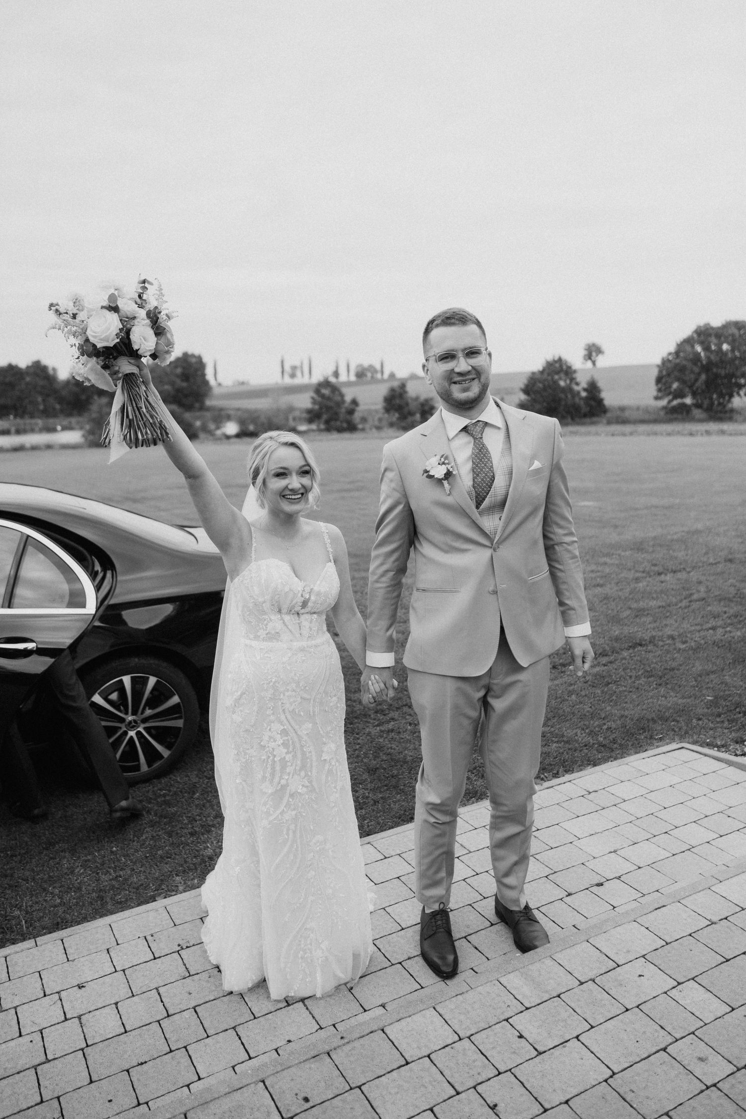 A joyful wedding couple poses outdoors on a paved walkway, celebrating their special day with enthusiastic gestures.
