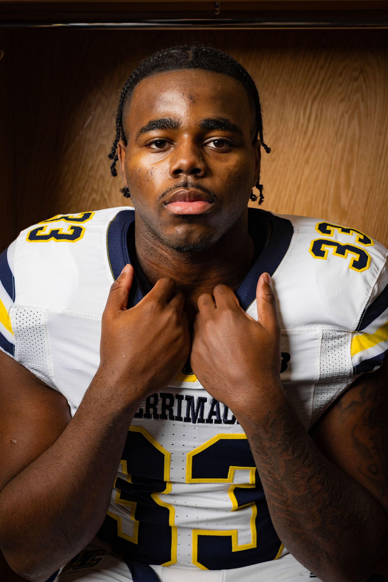 Football player wearing Merrimack #33 white and navy jersey poses in locker room for team portrait photo.