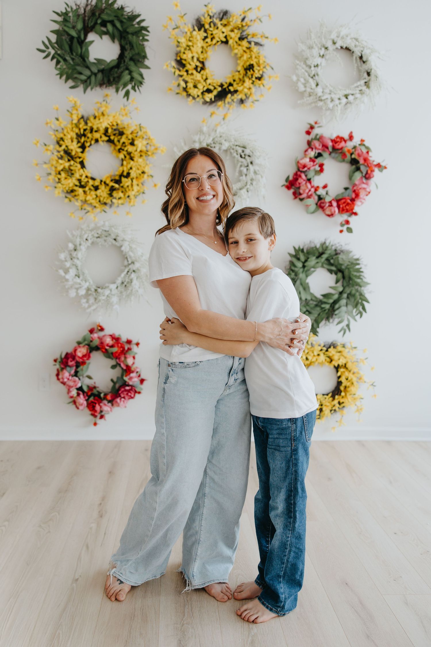 Mother and son in white tees and jeans posing before colorful floral wreath wall.