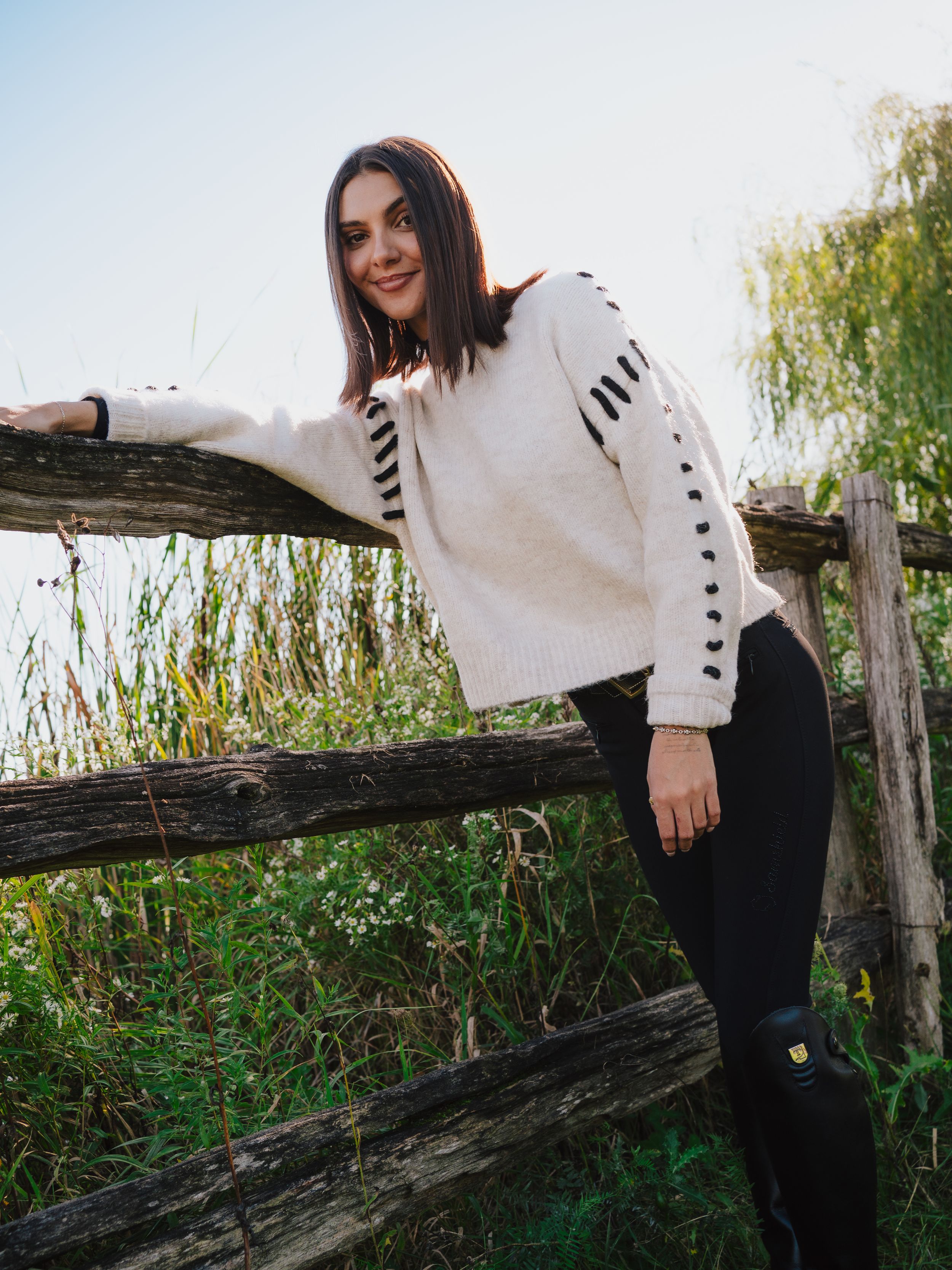 A person in a white sweater with black stripes leans against a rustic wooden fence surrounded by green foliage.