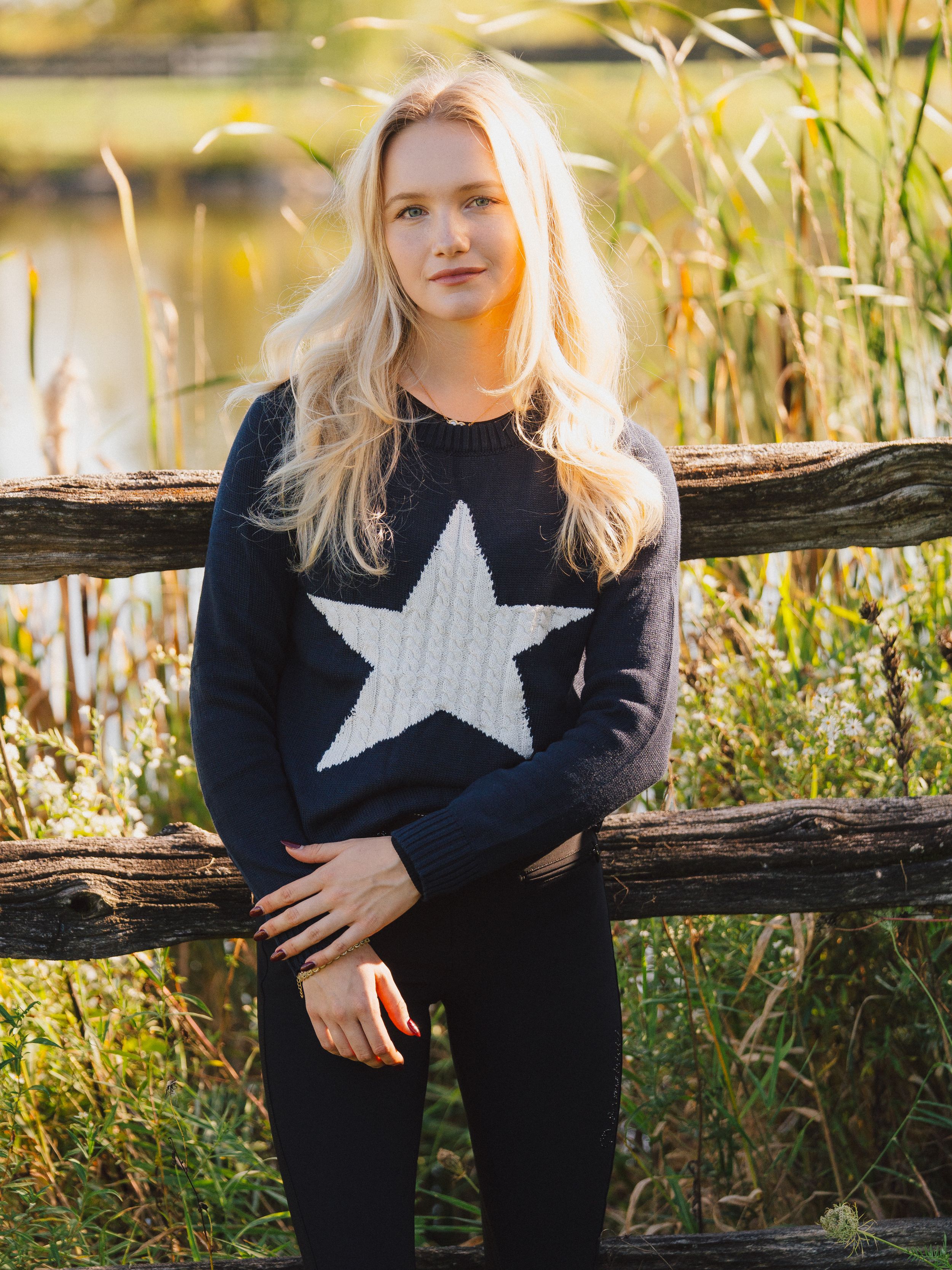 Person wearing navy sweater with white star design sitting by rustic wooden fence in golden autumn light.