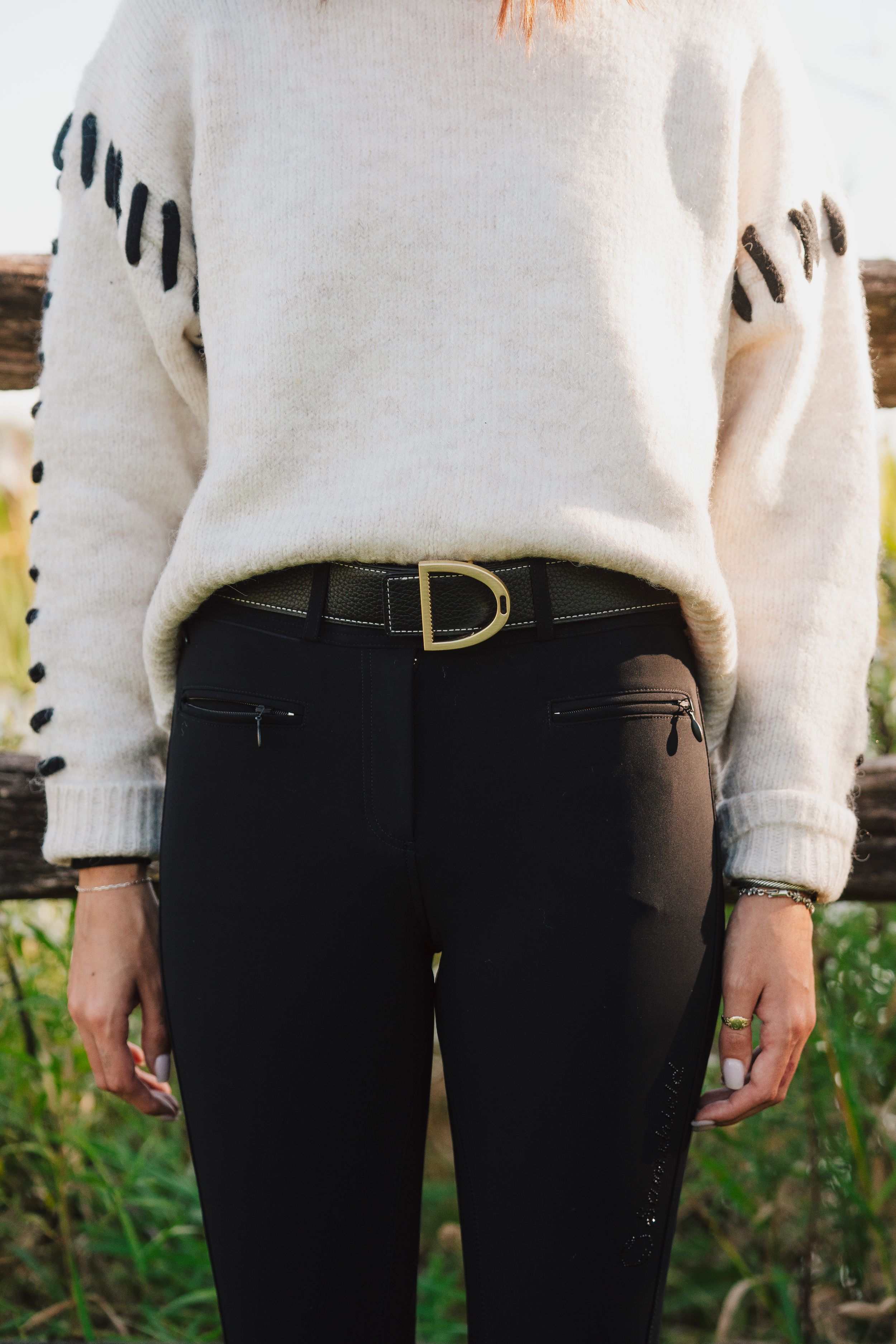 Close-up view of a person wearing a white sweater and black pants with a gold belt buckle detail.