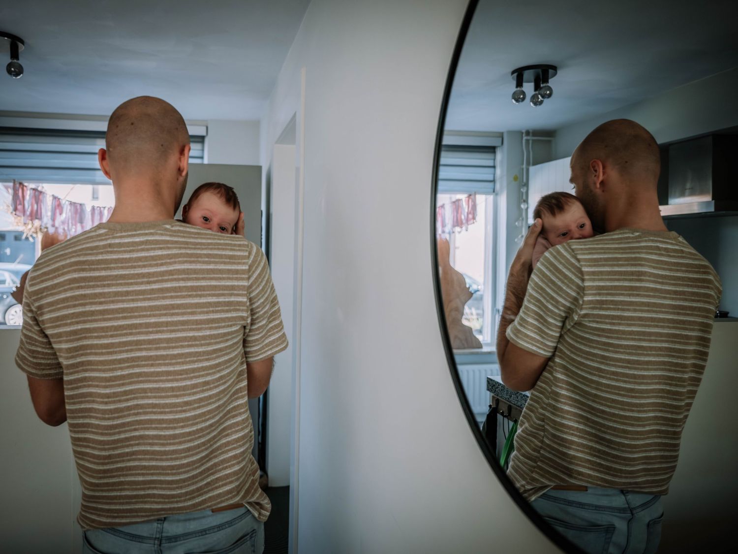 Bald man in striped t-shirt viewed from behind reflecting in round mirror in modern white room.