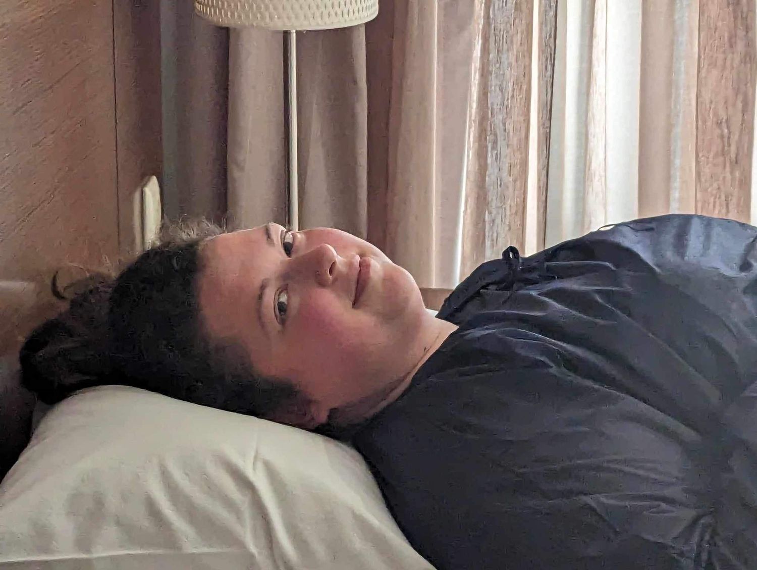 A person lying on a bed with white sheets and a curtained window in the background.