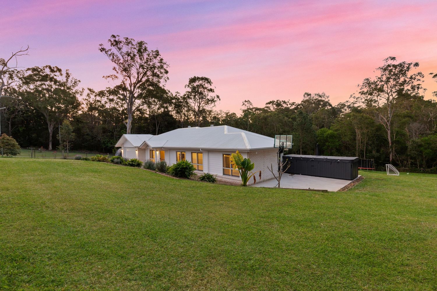 White country home surrounded by vast green paddock and tall eucalyptus trees under a vivid sunset sky.