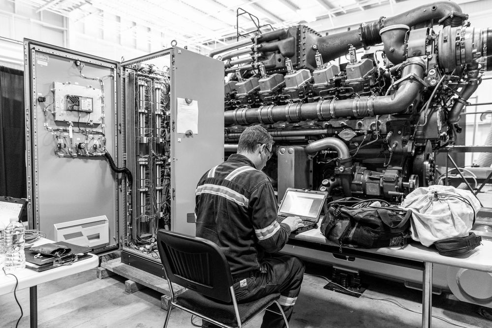 A technician works at a control panel next to large industrial engine machinery in black and white.