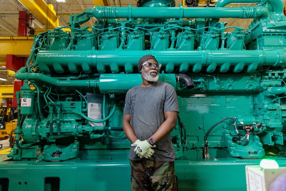 A mechanic works on a large turquoise diesel engine in an industrial maintenance facility.