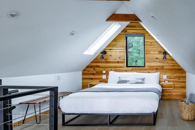 Cozy attic bedroom with wooden accent wall, white bedding, and a small window overlooking greenery.