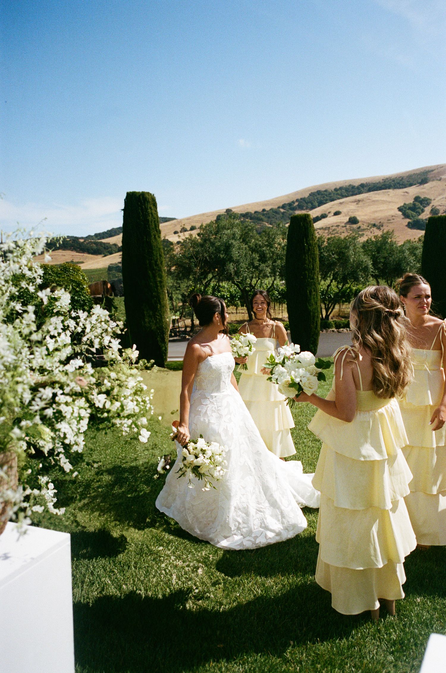 Wedding party in pale yellow dresses poses against a scenic hillside backdrop with cypress trees and white flowers.