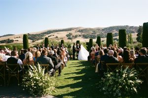 Panoramic view of an outdoor wedding ceremony with guests seated on a grassy hillside overlooking mountains.