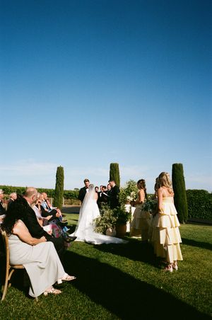 Wedding ceremony taking place outdoors on a grassy lawn with tall cypress trees under bright blue skies.