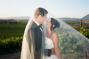 A couple shares a romantic kiss in a vineyard at sunset with rolling hills and grapevines in the background.