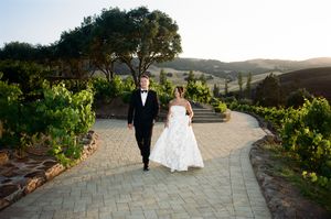 Newlyweds walk along a winding stone path surrounded by lush gardens and rolling hills at sunset.