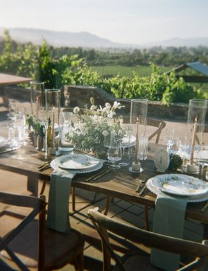 Elegant outdoor wedding reception table settings with rustic wooden tables and sage green linens overlooking a scenic vineyard.