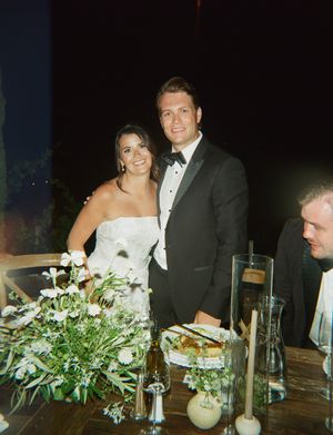 Wedding reception dinner table decorated with white floral centerpieces and glowing candles in evening lighting.