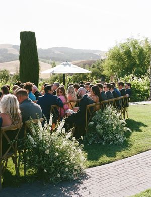 Wedding guests seated outdoors at a vineyard ceremony with mountain views and white chairs on a sunny day.